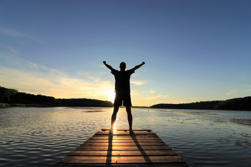 person on a wooden dock with arms outstretched looking over a lake