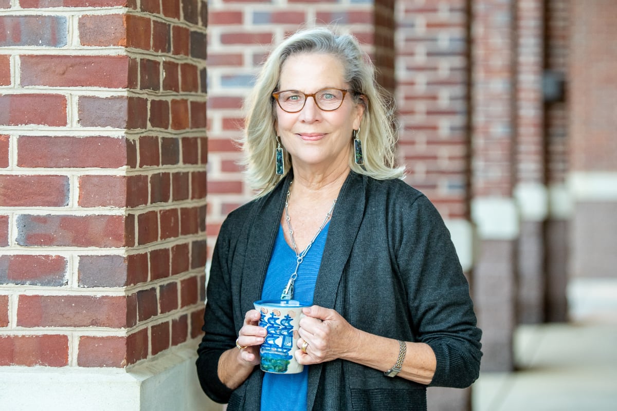 Jane O. Smith leaning on a brick column holding a mug