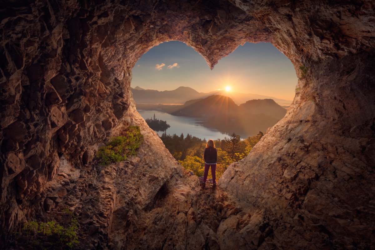 woman in a heart shaped rock formation looking over mountains and a lake at sunset or sunrise